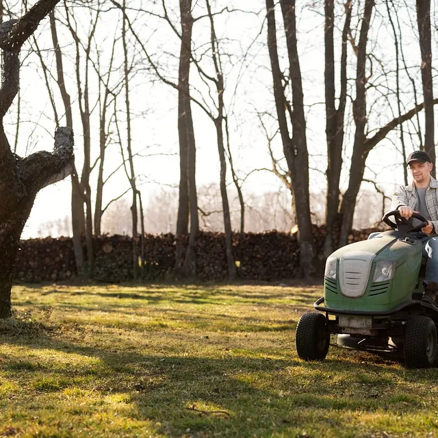 Smilende mann kjører plenklipper på gressplen omgitt av trær.